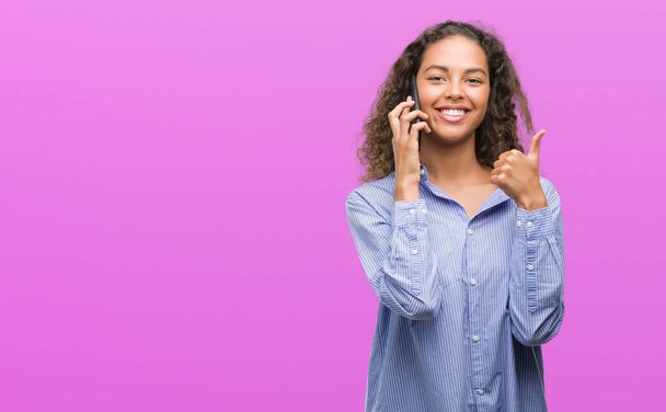 Young hispanic woman using smartphone happy with big smile doing ok sign, thumb up with fingers, excellent sign - Foto, Imagem