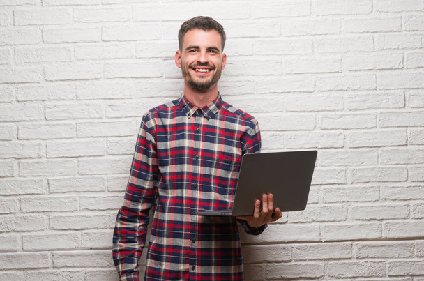 Молодий дорослий чоловік над цегляною стіною використовує ноутбук з щасливим обличчям, що стоїть і посміхається з впевненою посмішкою, показуючи зуби
 - Фото, зображення