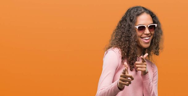 Belle jeune femme hispanique portant des lunettes de soleil pointant du doigt la caméra avec un visage heureux et drôle. Bonne énergie et vibes
. - Photo, image