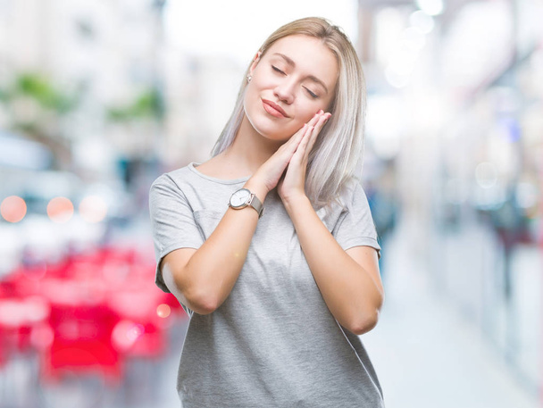 junge blonde Frau vor isoliertem Hintergrund schläft müde träumend und posiert mit den Händen zusammen, während sie mit geschlossenen Augen lächelt. - Foto, Bild