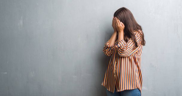 Młoda brunetka kobieta streszczenie szary mur z smutny wyrażenie obejmujące twarz rękami podczas płaczu. Koncepcja depresji. - Zdjęcie, obraz