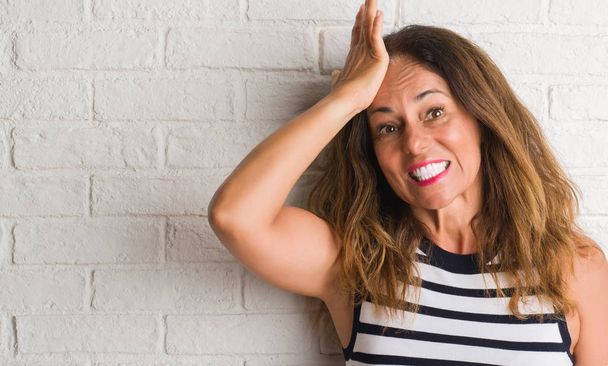 Middelbare leeftijd Latino vrouw over witte bakstenen muur met de hand op het hoofd benadrukt, geschokt met schaamte en verrassing gezicht, boos en gefrustreerd. Angst en boos voor fout. - Foto, afbeelding