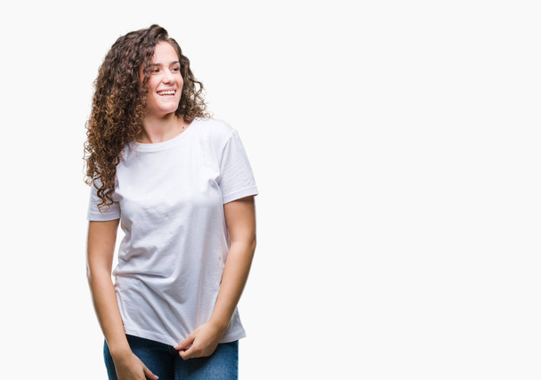 Beautiful brunette curly hair young girl wearing casual t-shirt over isolated background looking away to side with smile on face, natural expression. Laughing confident. - Photo, Image