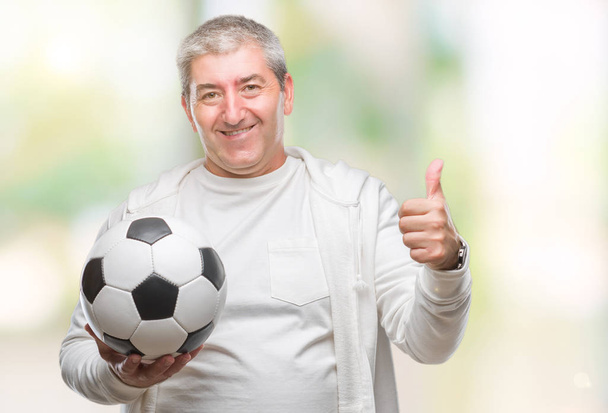 schöner älterer Mann hält Fußballball über isoliertem Hintergrund glücklich mit breitem Lächeln tut ok Zeichen, Daumen nach oben mit Fingern, ausgezeichnetes Zeichen - Foto, Bild