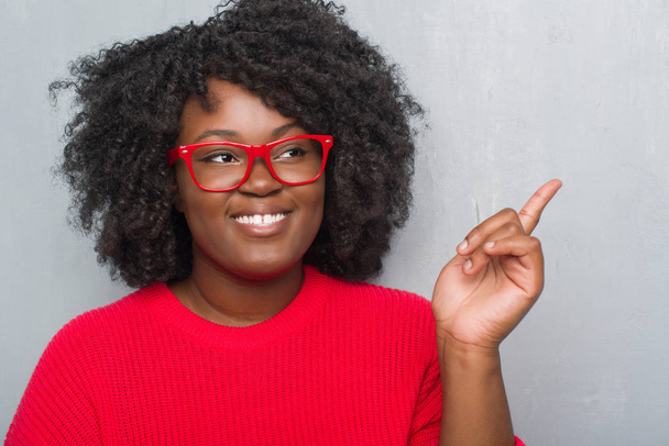 Jeune femme afro-américaine plus la taille sur le mur gris grunge portant un pull d'hiver avec un grand sourire sur le visage, pointant avec la main et le doigt vers le côté regardant la caméra
. - Photo, image