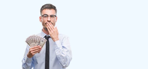 Young hipster business man holding dollars cover mouth with hand shocked with shame for mistake, expression of fear, scared in silence, secret concept - Photo, Image