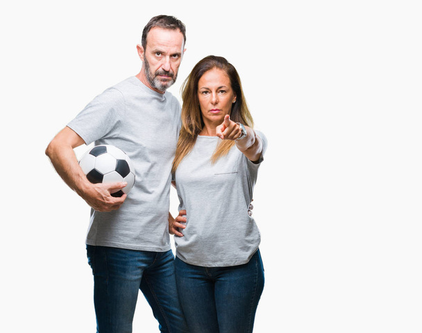 Orta yaş İspanyol çift futbol futbol topu tutan kamera ve size parmak ile işaret arka plan izole, el işareti, pozitif ve kendinden emin bir jest ön - Fotoğraf, Görsel