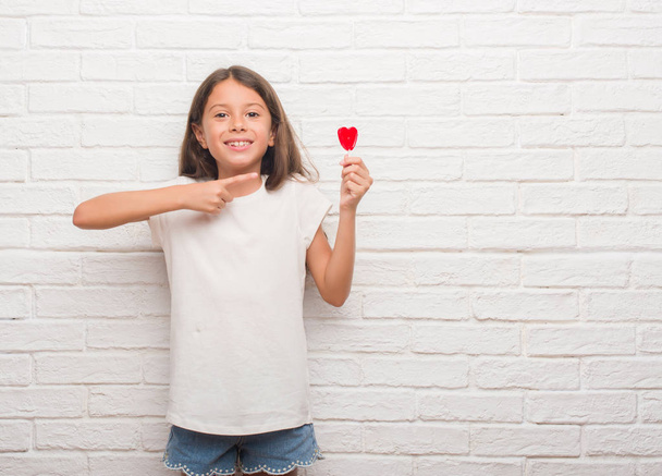 Jovem criança hispânica sobre parede de tijolo branco comendo doces pirulito coração vermelho muito feliz apontando com mão e dedo
 - Foto, Imagem