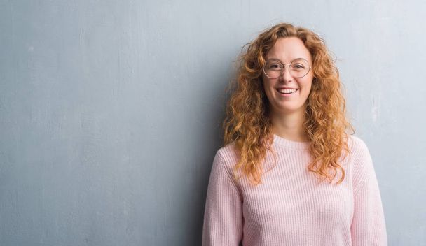 Young redhead woman over grey grunge wall wearing glasses with a happy face standing and smiling with a confident smile showing teeth - Foto, Imagem