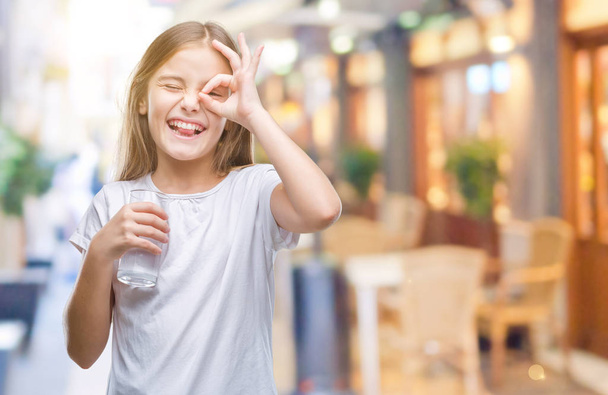 Young beautiful girl drinking glass of water over isolated background with happy face smiling doing ok sign with hand on eye looking through fingers - Φωτογραφία, εικόνα