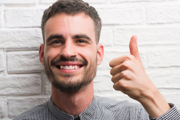 Jeune homme adulte debout sur un mur de briques blanches heureux avec un grand sourire faisant signe ok, pouce levé avec les doigts, excellent signe
 - Photo, image