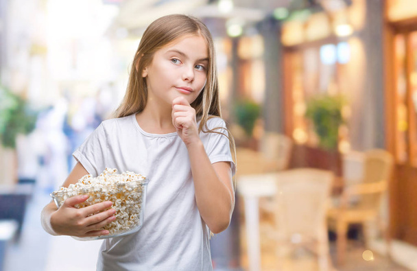 junge schöne Mädchen essen Popcorn-Snack isoliert Hintergrund ernste Gesicht denken über Frage, sehr verwirrte Idee - Foto, Bild