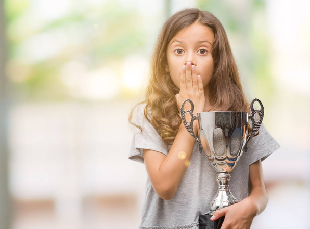 Brunette hispanic girl holding a trophy cover mouth with hand shocked with shame for mistake, expression of fear, scared in silence, secret concept - Photo, Image