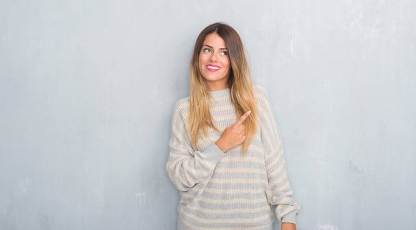 Jonge volwassen vrouw over grunge grijs muur dragen van winter trui met een grote glimlach op het gezicht, met de hand en de vinger te wijzen naar de kant te kijken naar de camera. - Foto, afbeelding