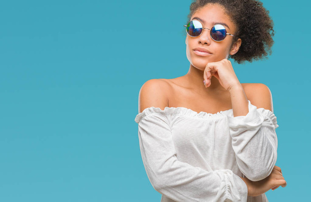 Молодая афро-американка в солнечных очках на изолированном фоне с рукой на подбородке думает о вопросе, задумчивом выражении лица. Улыбается с вдумчивым лицом. Концепция сомнений
. - Фото, изображение