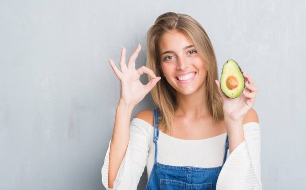 Красивая молодая женщина за гранж серые стены едят авокадо делает хорошо знак с пальцами, отличный символ
 - Фото, изображение