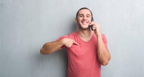 Mladý muž kavkazské přes zeď šedá grunge mluví po telefonu s překvapením tvář ukazováčkem na sebe - Fotografie, Obrázek