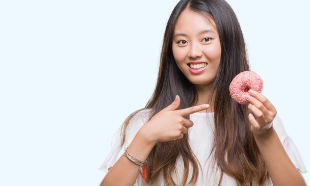 Young asian woman eating donut over isolated background very happy pointing with hand and finger - Foto, imagen