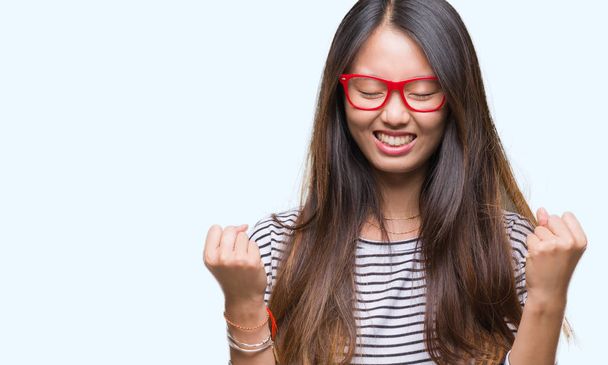 Young asian woman wearing glasses over isolated background very happy and excited doing winner gesture with arms raised, smiling and screaming for success. Celebration concept. - Φωτογραφία, εικόνα