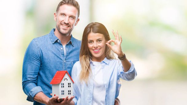 Young couple in love holding house over isolated background doing ok sign with fingers, excellent symbol - Foto, afbeelding