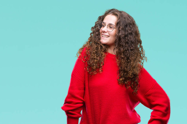 Beautiful brunette curly hair young girl wearing glasses and winter sweater over isolated background looking away to side with smile on face, natural expression. Laughing confident. - Valokuva, kuva