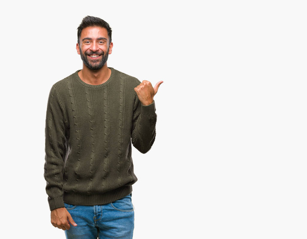 Ενηλίκων Ισπανόφωνος άνθρωπος φορώντας χειμώνα πουλόβερ πάνω από απομονωμένες φόντο χαμογελώντας με χαρούμενο πρόσωπο αναζητούν και τοποθετώντας το δείκτη στην πλευρά με τον αντίχειρα. - Φωτογραφία, εικόνα