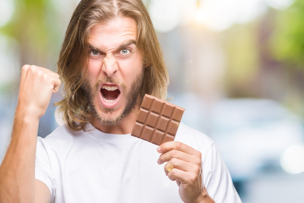 Młody przystojny mężczyzna z długimi włosami jedzenia czekolady na białym tle zirytowany i sfrustrowany krzycząc z gniewem, szalony i wrzask ręce, koncepcja gniew - Zdjęcie, obraz