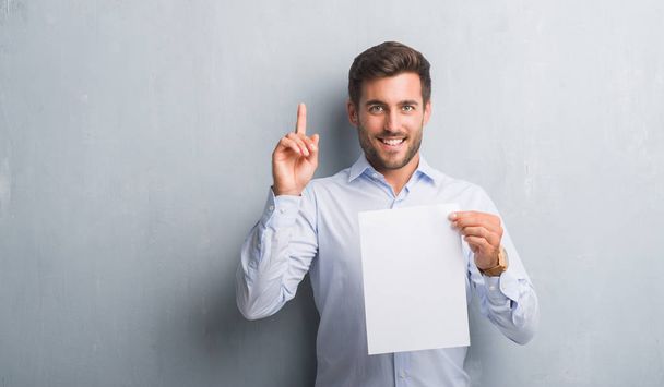 Pohledný mladý muž nad šedá grunge zeď hospodářství prázdný papír list smlouvy překvapilo s nápad nebo otázku ukazující prst s šťastné tváře, číslo jedna - Fotografie, Obrázek