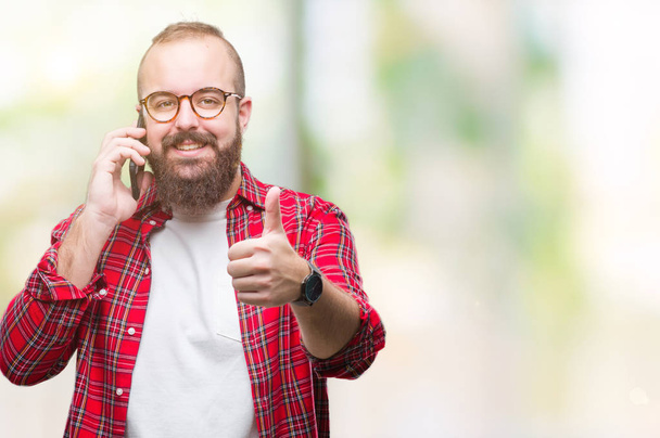 Jeune homme hipster caucasien parlant sur smartphone sur fond isolé heureux avec un grand sourire faisant signe ok, pouce levé avec les doigts, excellent signe
 - Photo, image