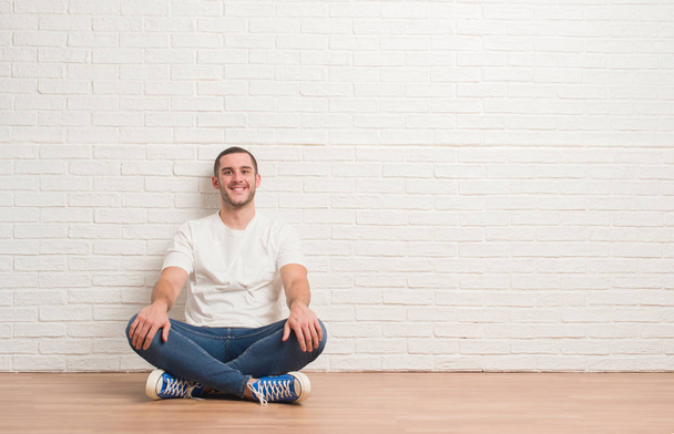 Young caucasian man sitting on the floor over white brick wall with a happy and cool smile on face. Lucky person. - Fotoğraf, Görsel
