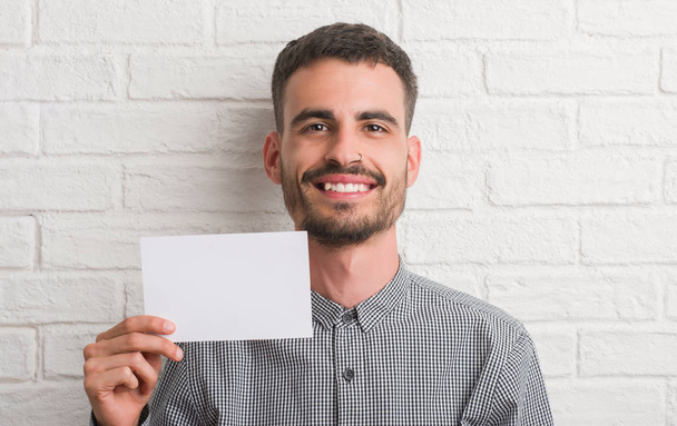 Νεαρός ενήλικος πάνω από τοίχο από τούβλα κρατώντας κενό χαρτί με ένα χαρούμενο πρόσωπο στέκονται και να χαμογελούν με αυτοπεποίθηση χαμόγελο δείχνει τα δόντια - Φωτογραφία, εικόνα