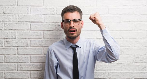 Young adult business man standing over white brick wall annoyed and frustrated shouting with anger, crazy and yelling with raised hand, anger concept - Photo, Image