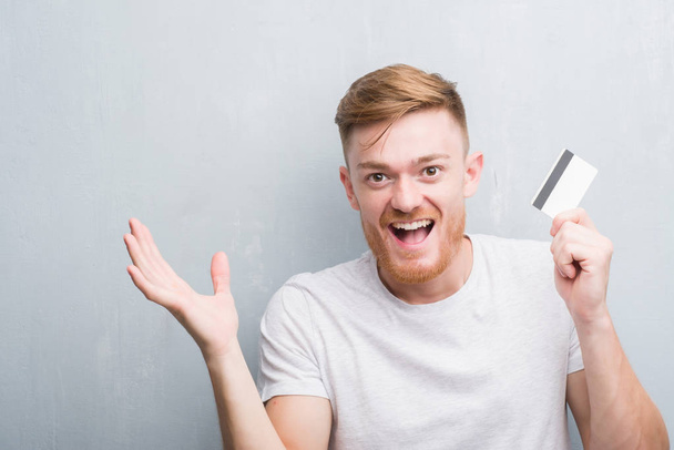 Young redhead man over grey grunge wall holding credit card very happy and excited, winner expression celebrating victory screaming with big smile and raised hands - Photo, Image