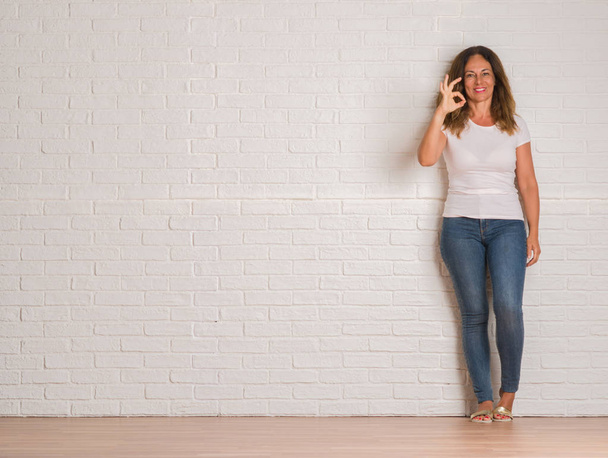 Middle age hispanic woman standing over white brick wall doing ok sign with fingers, excellent symbol - Foto, immagini