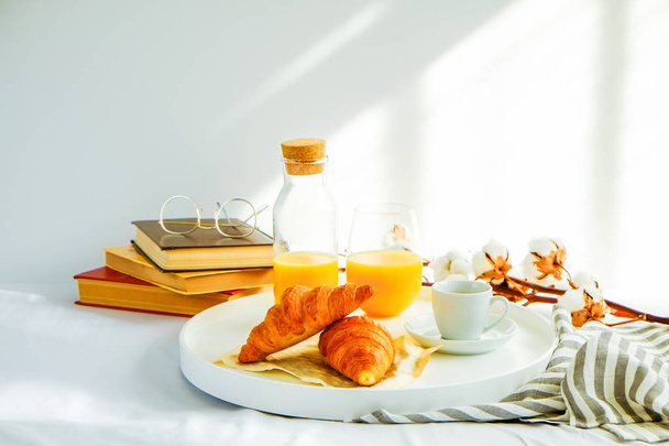 Ранковий сніданок у ліжку, білий піднос зі склянкою свіжого апельсинового соку, чашкою кави та французьким круасаном, старовинними окулярами та книгами, роздягненими ковдрами. Крупним планом, вид зверху, тло, простір для копіювання
. - Фото, зображення