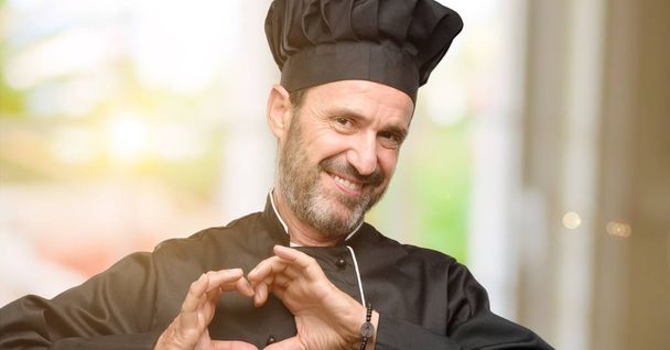 Senior cook man, wearing chef hat happy showing love with hands in heart shape expressing healthy and marriage symbol - Фото, изображение