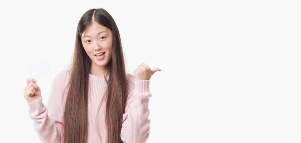 Jeune femme chinoise sur fond isolé tenant carte de visite pointant et montrant avec le pouce sur le côté avec sourire heureux visage
 - Photo, image