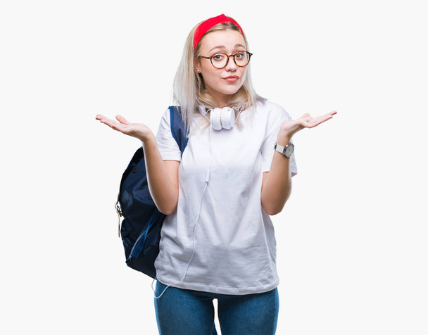 Młody student blond kobieta sobie okulary i plecak na na białym tle bladego pojęcia i mylić wyrażenie z ramiona i ręce podniesione. Koncepcja wątpliwości. - Zdjęcie, obraz