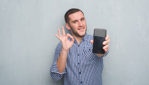 Giovane uomo caucasico sulla parete grigia grunge mostrando schermo bianco dello smartphone facendo segno ok con le dita, simbolo eccellente
 - Foto, immagini