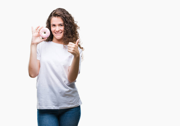 Young brunette girl eating donut over isolated background happy with big smile doing ok sign, thumb up with fingers, excellent sign - Zdjęcie, obraz