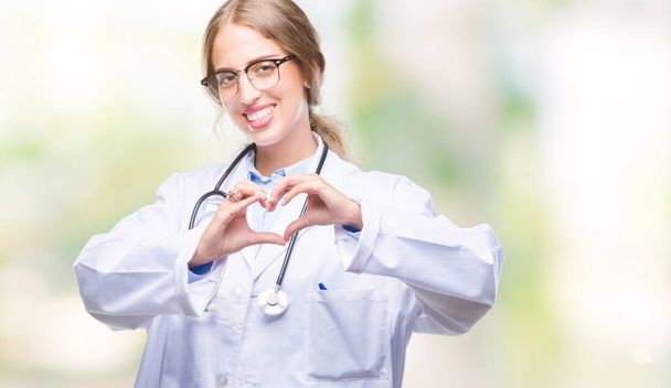 Beautiful young blonde doctor woman wearing medical uniform over isolated background smiling in love showing heart symbol and shape with hands. Romantic concept. - Photo, Image