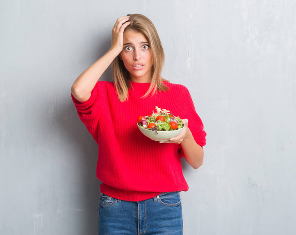 schöne junge Frau über grunge-grauer Wand beim Tomatensalat essen, gestresst mit der Hand auf dem Kopf, schockiert vor Scham und Überraschung, wütend und frustriert. Angst und Verärgerung wegen Fehlern. - Foto, Bild