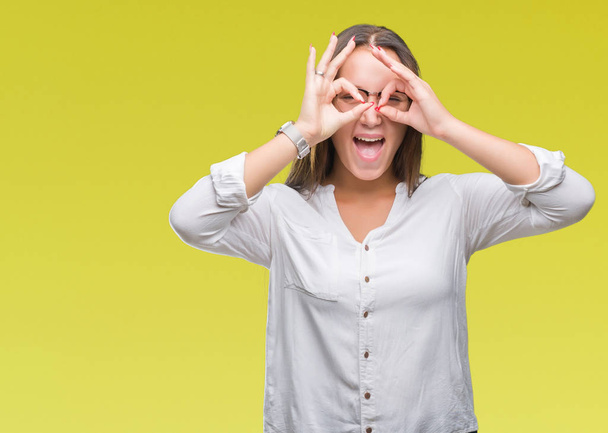 Young caucasian beautiful business woman wearing glasses over isolated background doing ok gesture like binoculars sticking tongue out, eyes looking through fingers. Crazy expression. - Foto, Imagen