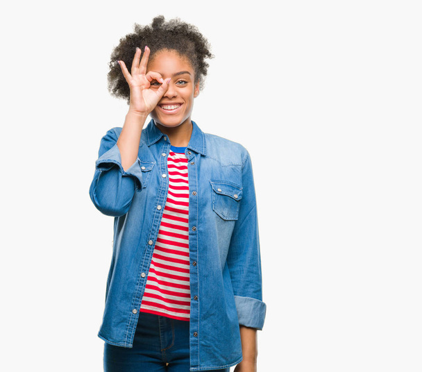 Young afro american woman over isolated background doing ok gesture with hand smiling, eye looking through fingers with happy face. - Valokuva, kuva