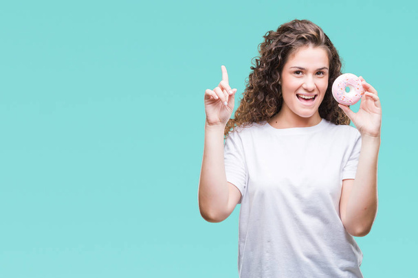 Young brunette girl eating donut over isolated background surprised with an idea or question pointing finger with happy face, number one - Foto, Imagen