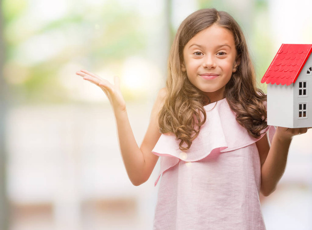 Brunette hispanic girl holding little house very happy and excited, winner expression celebrating victory screaming with big smile and raised hands - Photo, Image