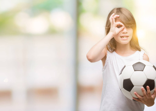 Młoda Blondynka malucha trzymając piłki nożnej z szczęśliwy twarz uśmiechający się robi ok znak ręką na oko patrząc przez palce - Zdjęcie, obraz