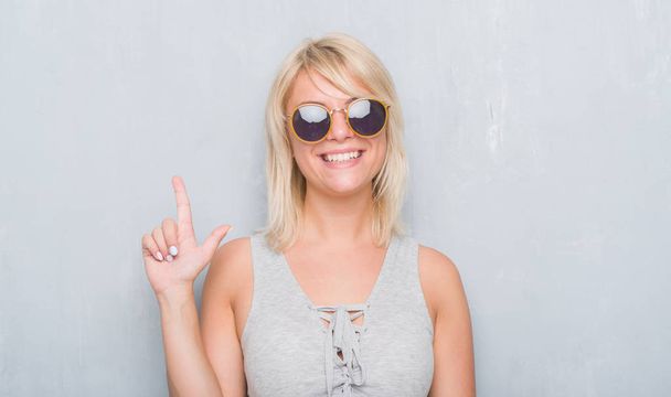Aikuinen valkoihoinen nainen yli grunge harmaa seinä yllään retro aurinkolasit yllättynyt idea tai kysymys osoittaa sormella onnellinen kasvot, numero yksi
 - Valokuva, kuva