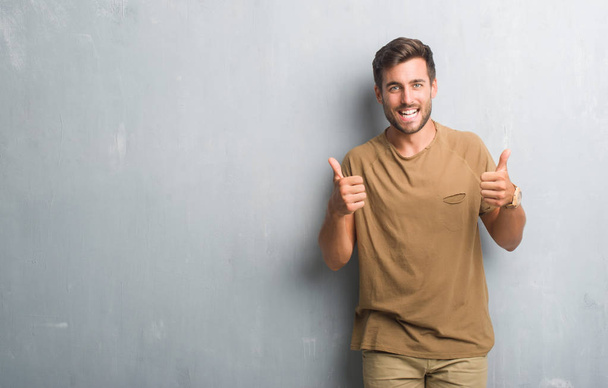 Handsome young man over grey grunge wall success sign doing positive gesture with hand, thumbs up smiling and happy. Looking at the camera with cheerful expression, winner gesture. - Photo, Image