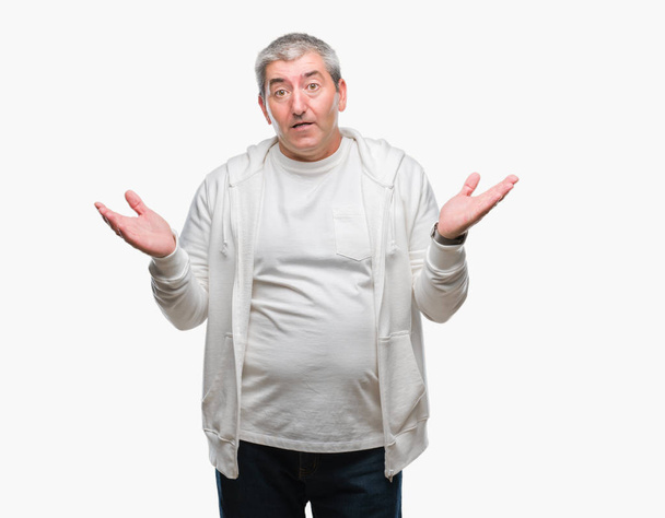 Pěkný starší muž nosí sportovní oblečení nad izolované pozadí bezradný a zmateni výraz se zbraněmi a rukama zdviženýma. Pochybnost koncept. - Fotografie, Obrázek